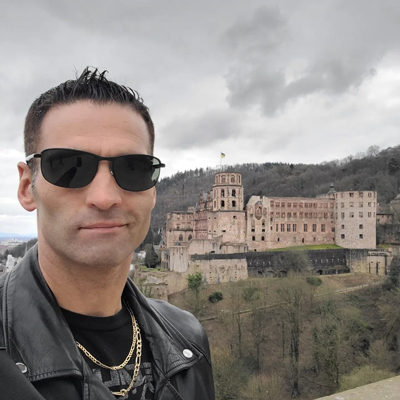 Man in sunglasses and leather jacket posing before a historic castle on a cloudy day.