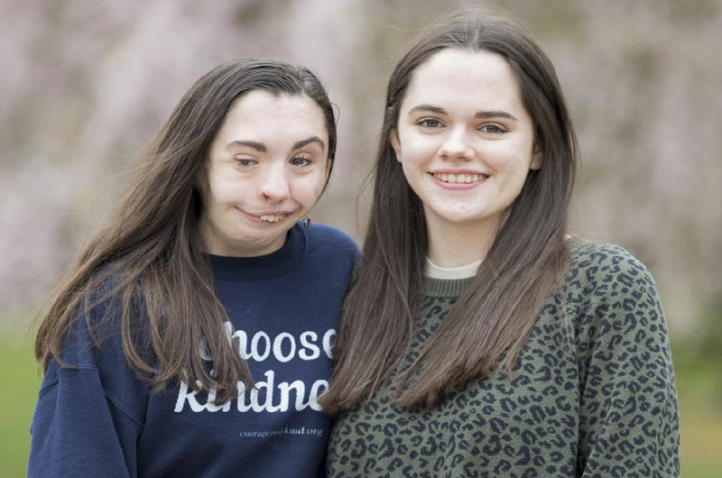 Twin sisters, including one wearing a blue sweatshirt that says Choose Kindness, are standing in front of a blurred background.