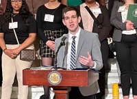 male student in grey suit talking at podium with other students behind him