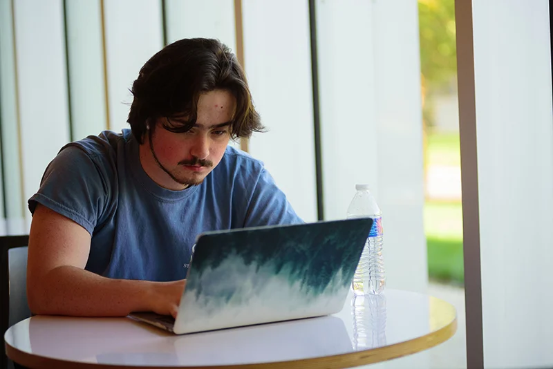 man sitting at table near window with laptop and water bottle The Future of Online Learning: Why Marywood’s Graduate Programs Stand Out