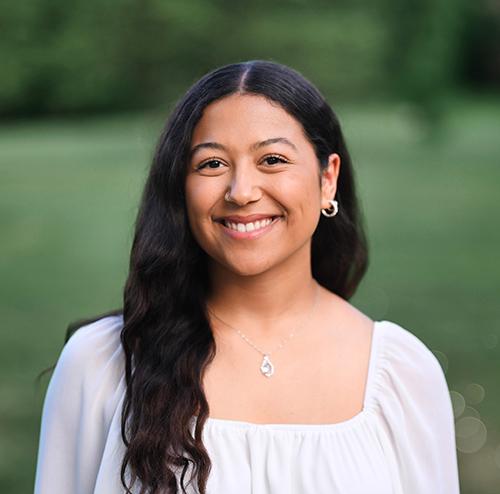 girl with long black hair in grass field smiling wearing a white shirt and silver necklace