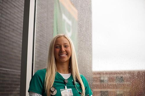 A smiling woman in green scrubs with a stethoscope by a rain-speckled window.