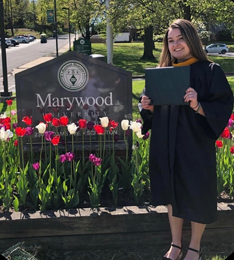 A female student in a graduation cap and gown, holding a diploma, standing next to a marble sign that reads 