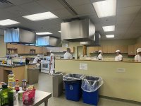 kitchen area with ingredients on the table and students in white scrubs and hats talking to eachother