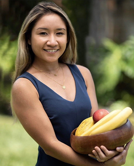 A smiling woman with shoulder-length hair, wearing a sleeveless navy blue dress, holding a wooden bowl filled with assorted fruits.
