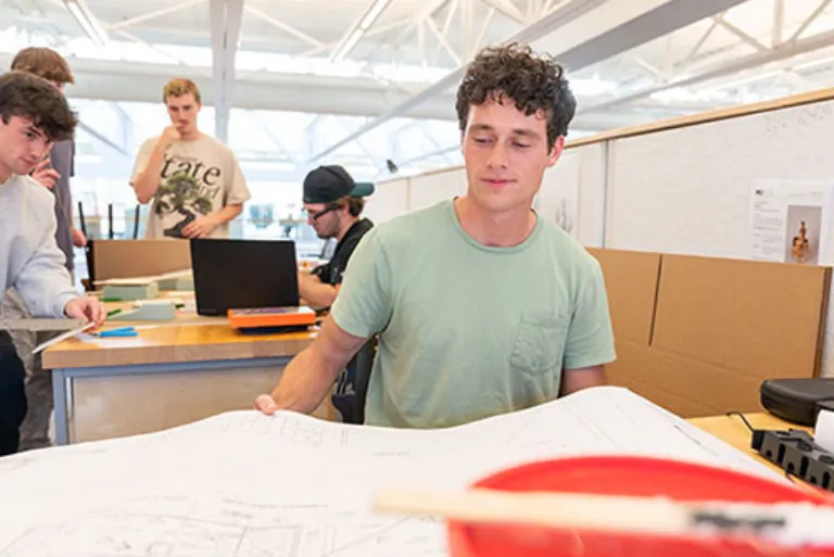 guy sitting at desk looking at blueprints with students behind him working on their own projects