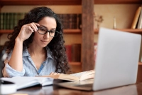 woman with glasses and long curly black hair sitting at desk to study with laptop and books open