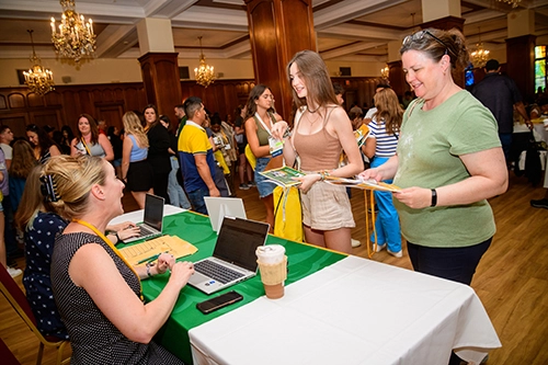 a mom and daughter standing up in front of a table talking to a woman sitting at the table behind her laptop